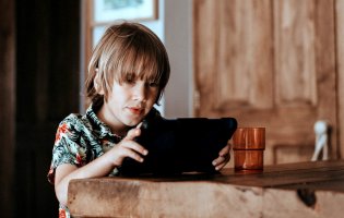 Ein Kind mit einem bunten Hemd hält ein Tablet am Holztisch und schaut ganz vertieft.