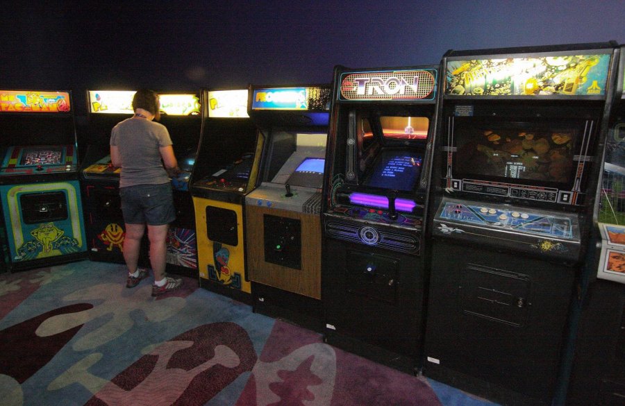 Zu sehen ist eine Videospielhalle mit verschiedenen Automaten. Die Spiele Miss Pacman und Tron sind zu erkennen. Vor einem der Automaten steht eine Person und spielt.