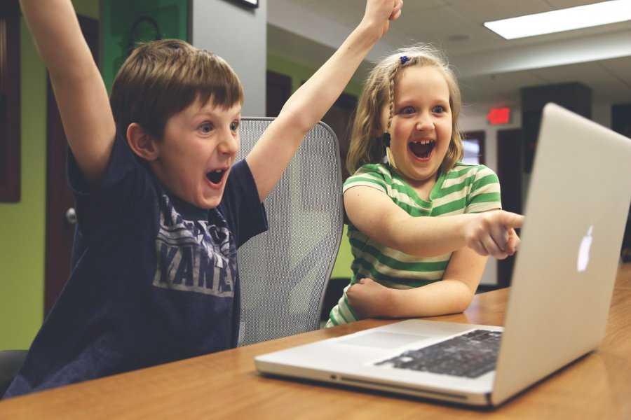 Zwei Kinder sitzen vor einem Laptop und spielen daran. Das Mädchen zeigt auch den Bildschirm, während der Kunge seine Arme nach oben reißt und jubelt. Beide lachen und sehen glücklich aus.
