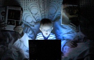 Ein kleines Kind sitzt zwischen den Eltern im Bett hinter einem leuchteten Bildschirm. Die Eltern sind hinter Zeitschriften verdeckt, die sie lesen.